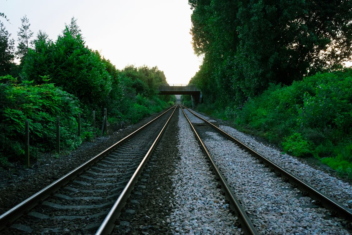A double railway track running through green countryside under a footbridge, like the line that serves the Stansted airport terminal