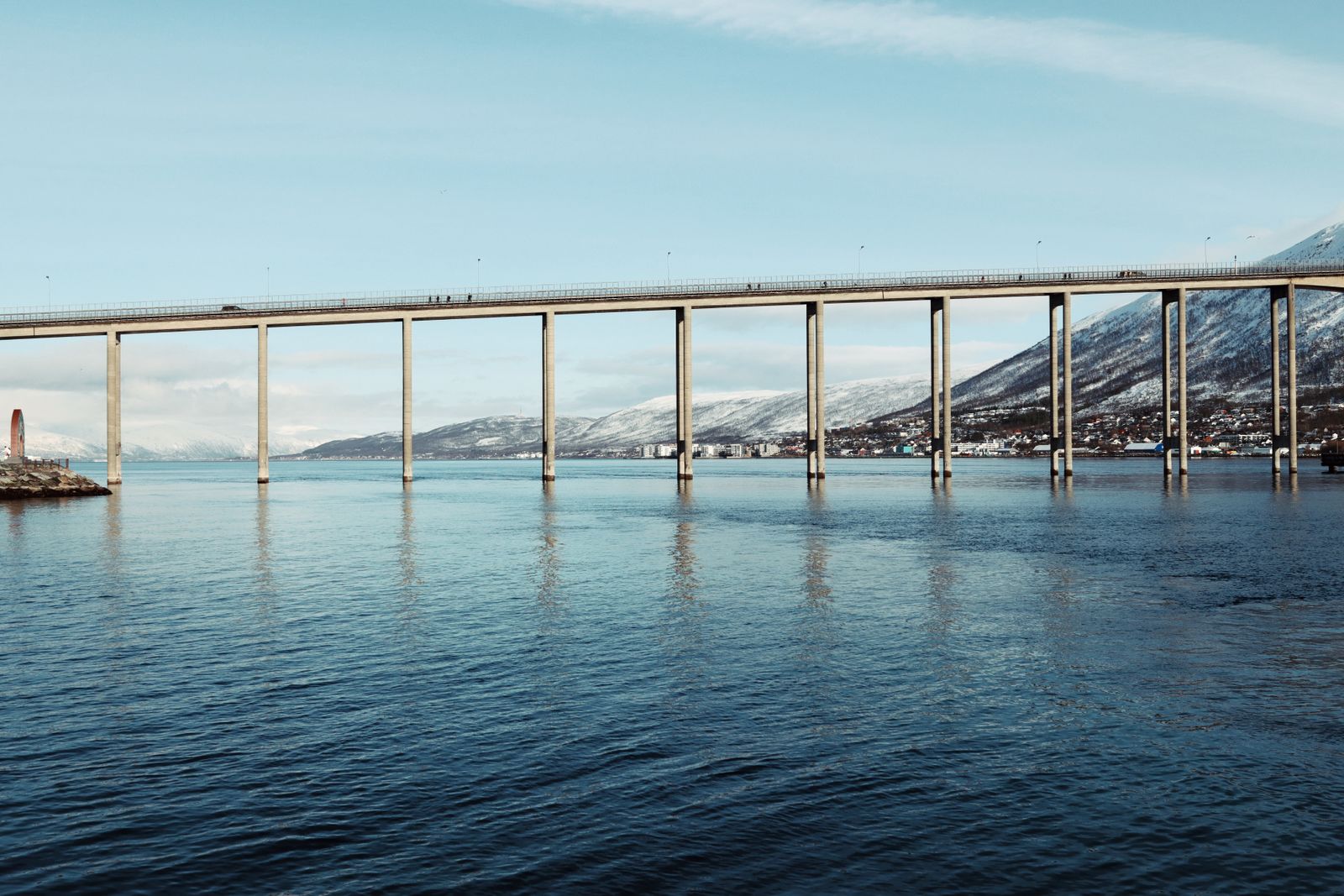 The long Tromso Bridge on concrete pillars over calm blue water with snowy hills behind