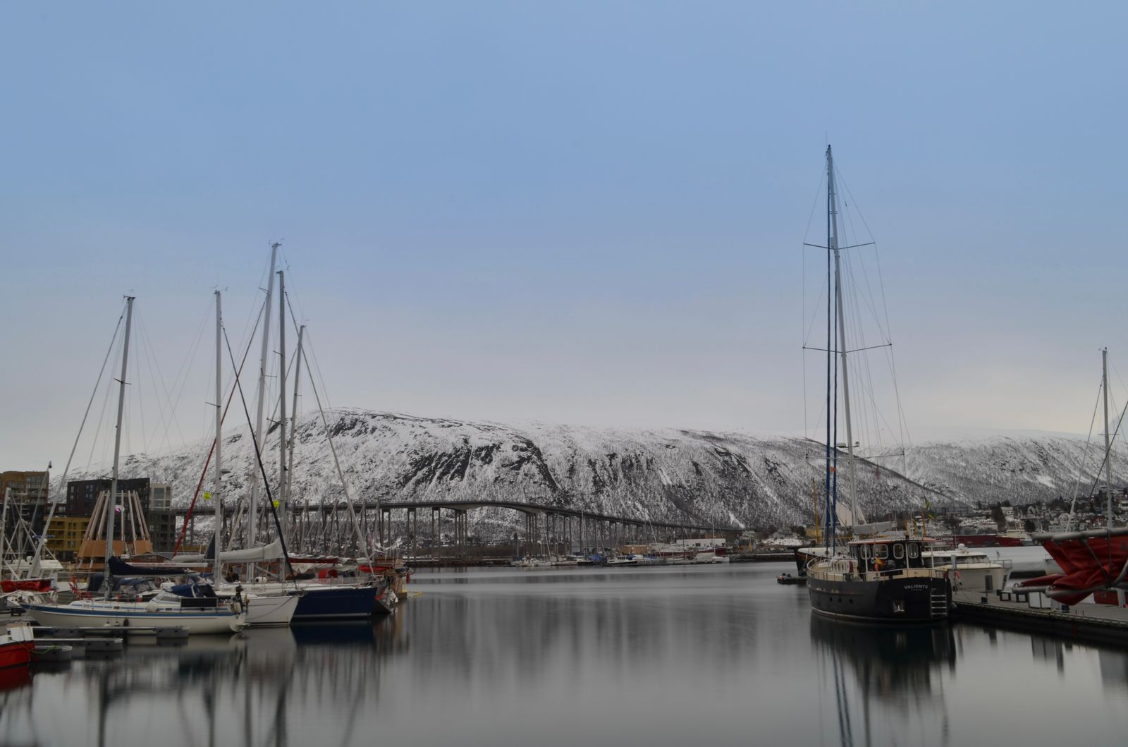 Moored sailing boats in Tromso harbour with the Tromso Bridge and snow-covered mountain behind