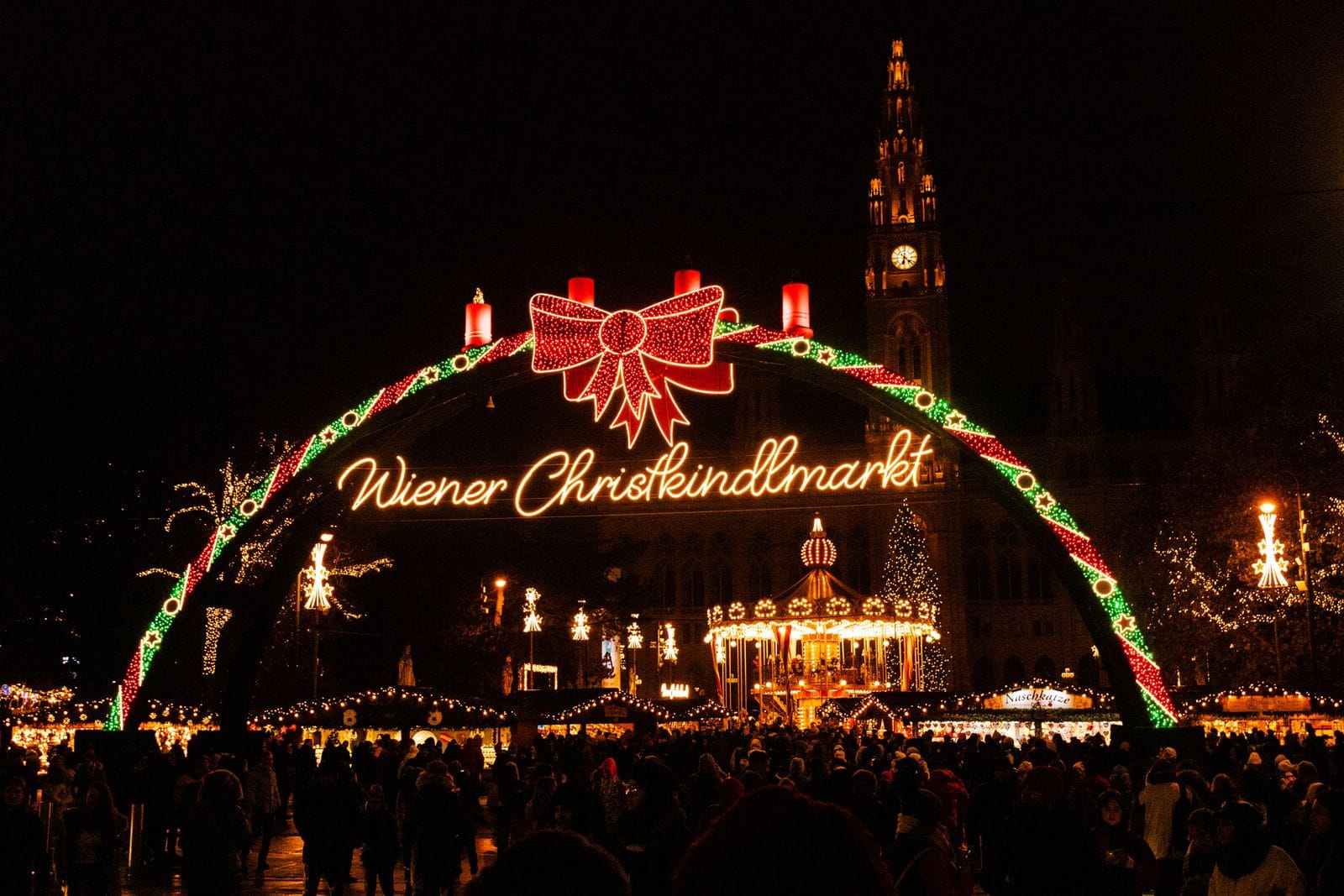 The illuminated Wiener Christkindlmarkt arch in front of Vienna City Hall at night, with crowds below